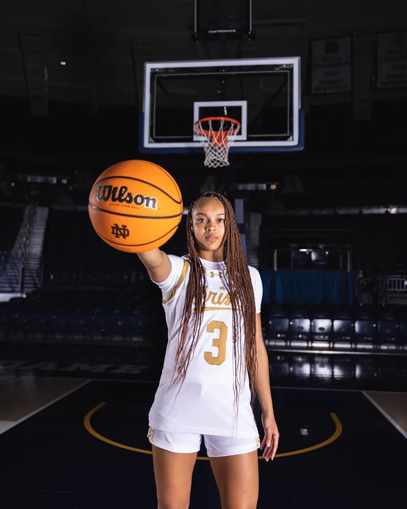 Hannah Hidalgo in Notre Dame's white number 3 uniform, holding a basketball on an empty court