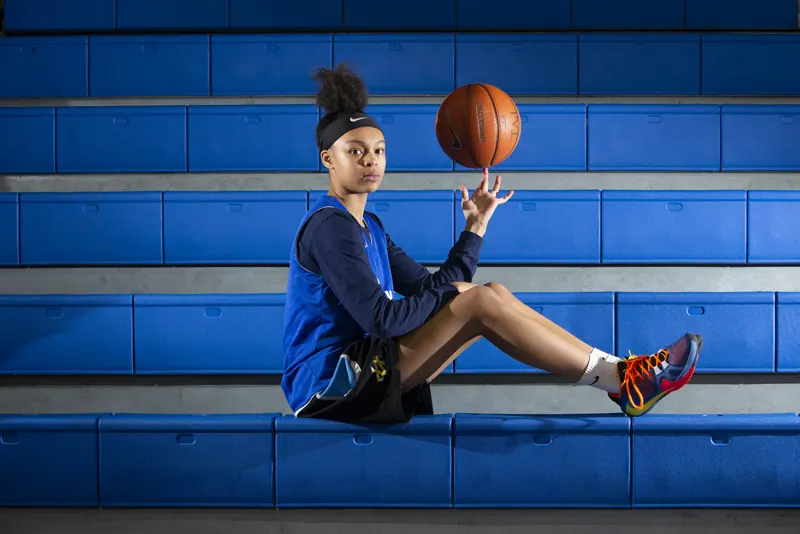 A teenage Hannah Hidalgo sits on blue bleachers, spinning a basketball on one finger